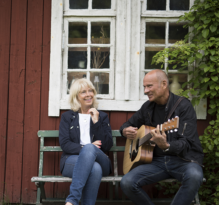 Lasse Holm jouant de la guitare dehors avec une femme devant un chalet rouge.