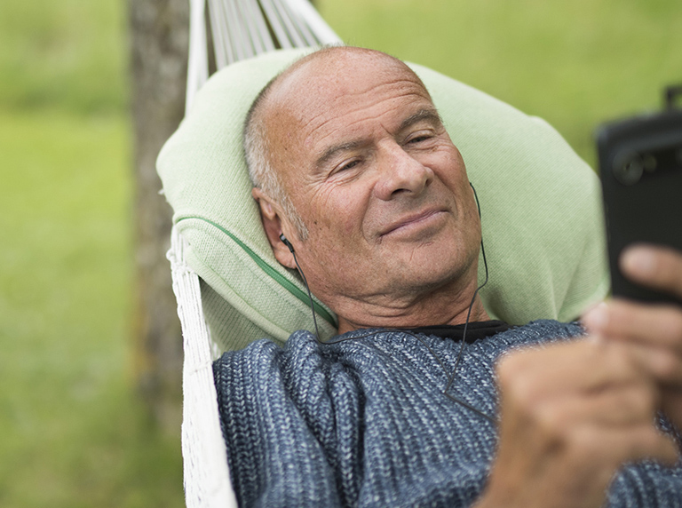 Older man relaxing in a hammock using a smartphone and listening with earphones