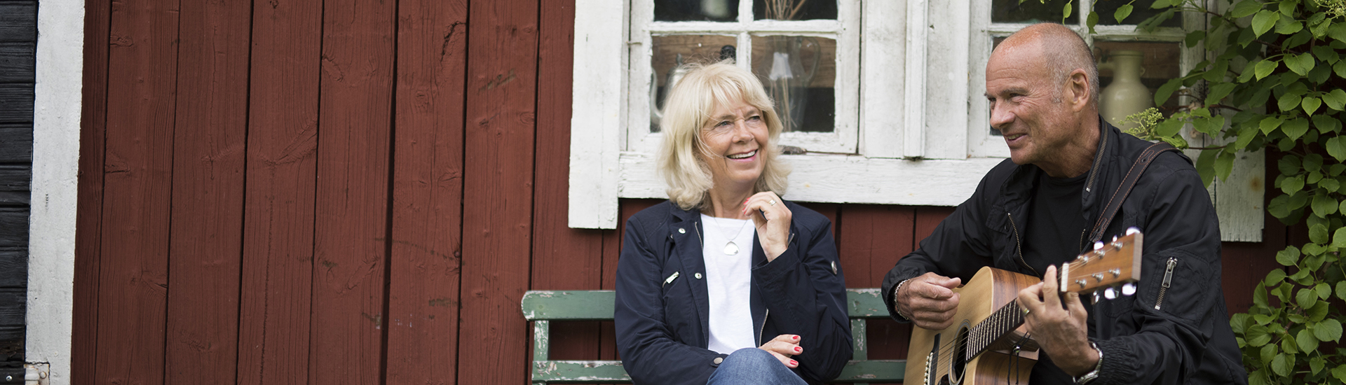 Lasse Holm playing guitar with woman outside a red cottage.